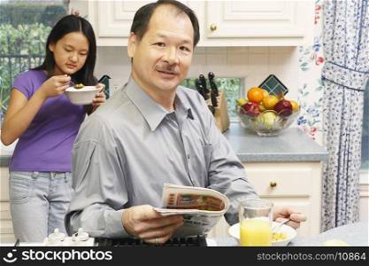 Portrait of a father holding a newspaper with his daughter eating breakfast behind him
