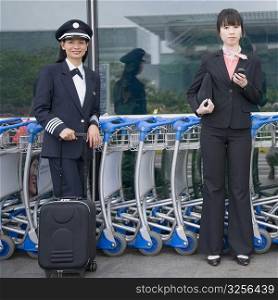Portrait of a female cabin crew holding a mobile phone and standing with a female pilot at an airport