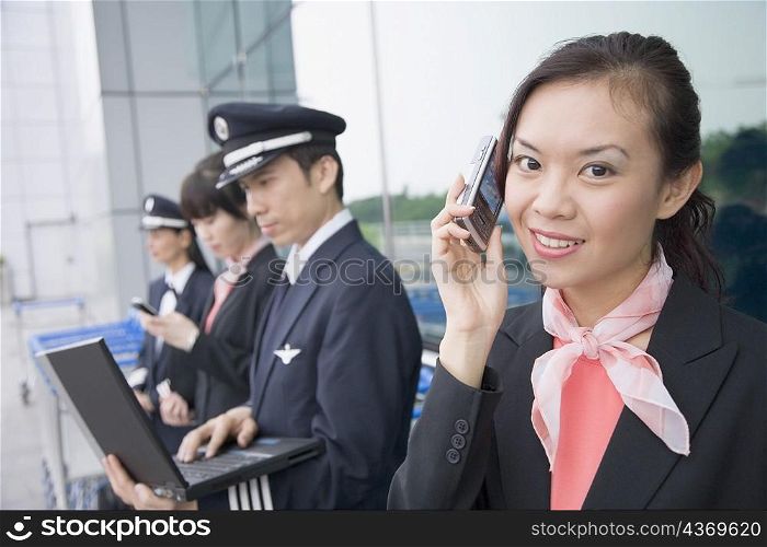 Portrait of a female cabin crew talking on a mobile phone with a pilot using a laptop in the background