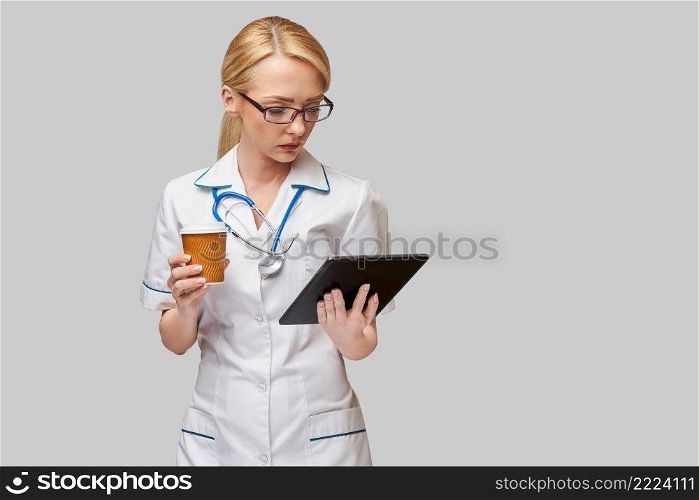 Portrait of a female doctor holding paper cup of coffee and holding tablet pc pad standing over grey background.. Portrait of a female doctor holding paper cup of coffee and holding tablet pc pad standing over grey background