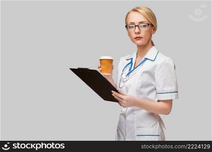 Portrait of a female doctor holding paper cup of coffee and holding tablet pc pad standing over grey background.. Portrait of a female doctor holding paper cup of coffee and holding tablet pc pad standing over grey background