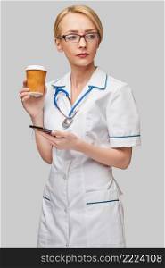 Portrait of a female doctor holding paper cup of coffee and talking on the phone standing over grey background.. Portrait of a female doctor holding paper cup of coffee and talking on the phone standing over grey background
