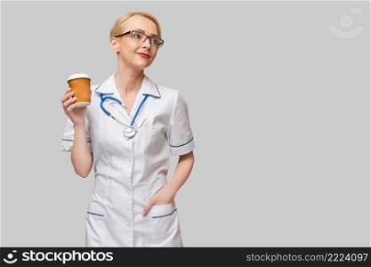 Portrait of a female doctor holding paper cup of coffee standing over grey background.. Portrait of a female doctor holding paper cup of coffee standing over grey background