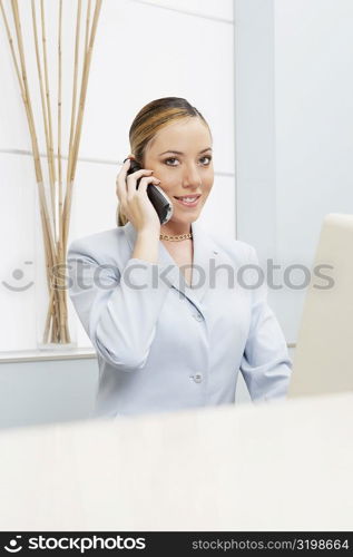 Portrait of a female receptionist talking on a mobile phone in front of a computer