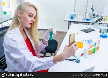 portrait of a female researcher carrying out research in a chemistry lab scientist holding test tube with sample taking a selfie with focus to smartphone in Laboratory analysis background