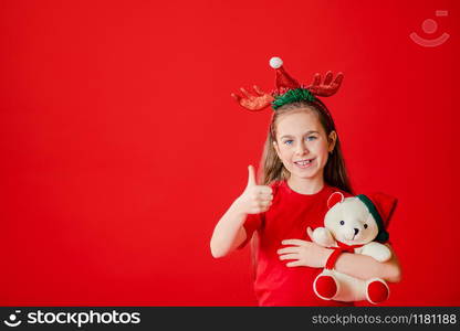 Portrait of a funny cheerful girl who is wearing hristmas pajama and a bandage of horns on her head. Girl is hugging a teddy bear. Girl isolated on a bright red background, copy space.. Portrait of a funny cheerful girl who is wearing hristmas pajama and a bandage of horns on her head. Girl is hugging a teddy bear.