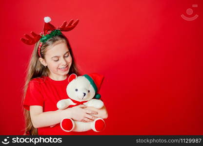 Portrait of a funny cheerful girl with a bandage of horns on her head hugging a teddy bear in Christmas pajamas isolated on a bright red background. The child points a hand, a place for text.. Portrait of a funny cheerful girl with a bandage of horns on her head hugging a teddy bear in Christmas pajamas isolated on a bright red background.