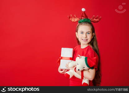 Portrait of a funny cheerful girl with a bandage of horns on her head hugging a teddy bear in Christmas pajamas isolated on a bright red background. The child points a hand, a place for text.. Portrait of a funny cheerful girl with a bandage of horns on her head hugging a teddy bear in Christmas pajamas isolated on a bright red background.