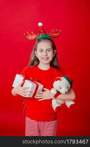 Portrait of a funny cheerful girl with a bandage of horns on her head hugging a teddy bear in Christmas pajamas isolated on a bright red background. The child points a hand, a place for text.. Portrait of a funny cheerful girl with a bandage of horns on her head hugging a teddy bear in Christmas pajamas isolated on a bright red background.