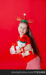 Portrait of a funny cheerful girl with a bandage of horns on her head hugging a teddy bear in Christmas pajamas isolated on a bright red background. The child points a hand, a place for text.. Portrait of a funny cheerful girl with a bandage of horns on her head hugging a teddy bear in Christmas pajamas isolated on a bright red background.