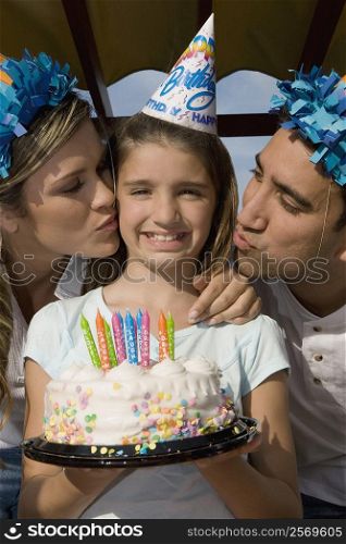 Portrait of a girl holding a birthday cake and her parents kissing her