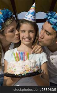 Portrait of a girl holding a birthday cake and her parents kissing her