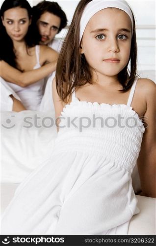 Portrait of a girl reclining on the bed with her parents sitting behind her