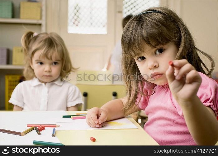 Portrait of a girl showing a crayon with her friend sitting in the background