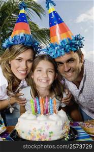 Portrait of a girl with her parents standing in front of a birthday cake and smiling
