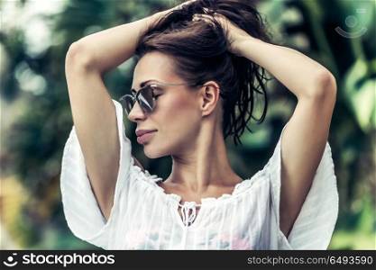 Portrait of a gorgeous female wearing stylish sunglasses and with pleasure touching her hair posing over exotic plants background, photoshoot on the tropical beach resort, enjoying summer vacation. Gorgeous female portrait