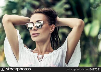 Portrait of a gorgeous female wearing stylish sunglasses posing over exotic plants background, photoshoot on the beach resort, enjoying summer vacation. Gorgeous female on the tropical resort