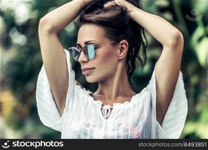 Portrait of a gorgeous female wearing stylish sunglasses posing over exotic plants background, photoshoot on the luxury beach resort, enjoying summer vacation. Gorgeous female on the tropical resort