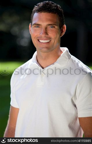 Portrait of a handsome man in his thirties wearing a white polo shirt outside in a sunlit green field.