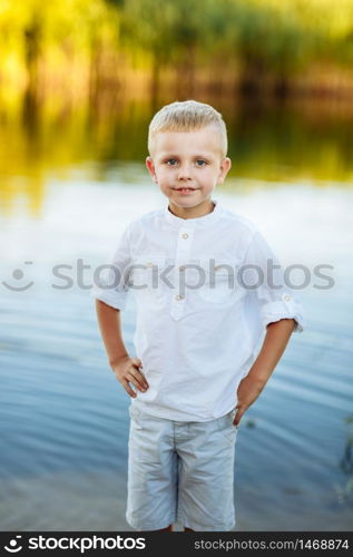 Portrait of a happy and beautiful little boy with blond hair and a white shirt, Happy childhood. Positive emotion. portrait near the river in nature.. Portrait of a happy and beautiful little boy with blond hair and a white shirt, Happy childhood. Positive emotion. portrait near the river in nature