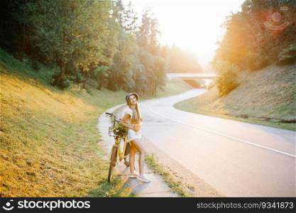 Portrait of a happy beautiful young girl with a bicycle and flowers on the background of a forest by the road in the sunlight in the open air