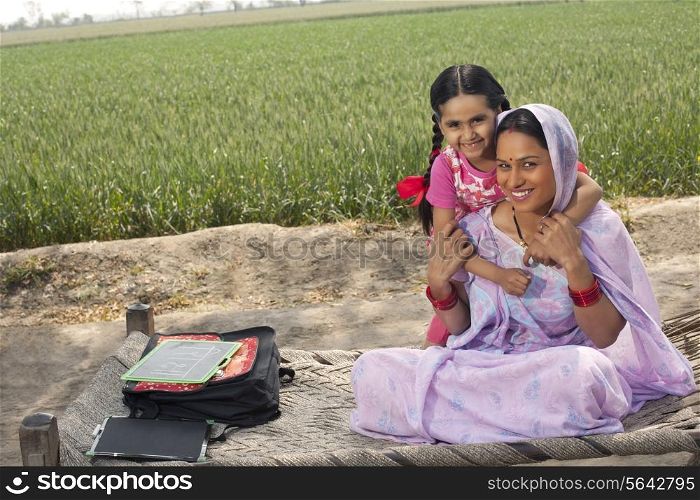 Portrait of a happy Indian mother and daughter sitting on cot with field in background