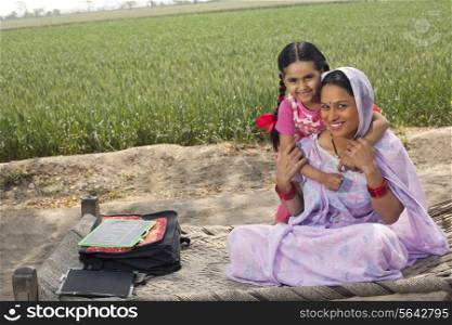 Portrait of a happy Indian mother and daughter sitting on cot with field in background
