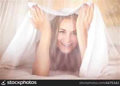 Portrait of a happy joyful woman in the bed, playing and having fun at home, peeking out from under the sheets and looking through the tulle