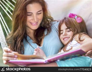 Portrait of a Happy Little Girl with Mother in the Hammock Reading Book. With Pleasure Spending Sunny Day on Backyard Together. Happy Summer Holidays.. Happy Family Outdoors