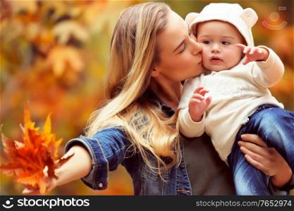 Portrait of a happy mother holding her precious little son, kissing him and showing to him dry maple leaves, having fun in the autumn park