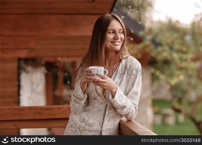 Portrait of a happy smiling young woman in stylish nightwear with a cup of coffee or tea meeting the day standing on the hotel terrace with a gorgeous view of the mountains. place for inscription