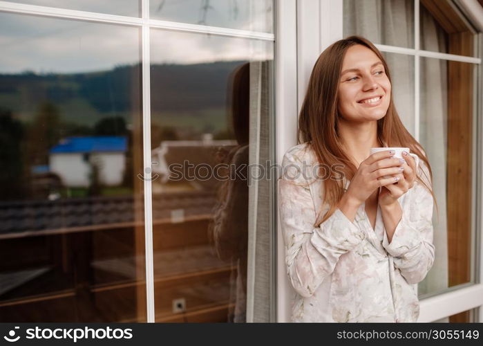 Portrait of a happy smiling young woman in stylish nightwear with a cup of coffee or tea meeting the day standing on the hotel terrace with a gorgeous view of the mountains. place for inscription