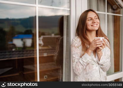 Portrait of a happy smiling young woman in stylish nightwear with a cup of coffee or tea meeting the day standing on the hotel terrace with a gorgeous view of the mountains. place for inscription