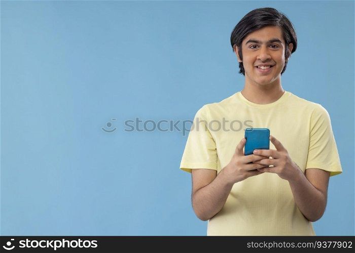 Portrait of a happy teenage boy using Smartphone while standing against blue background