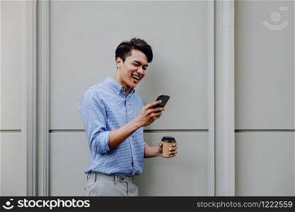 Portrait of a Happy Young Businessman Using Mobile Phone. Lifestyle of Modern People. Standing by the Wall with Coffee Cup