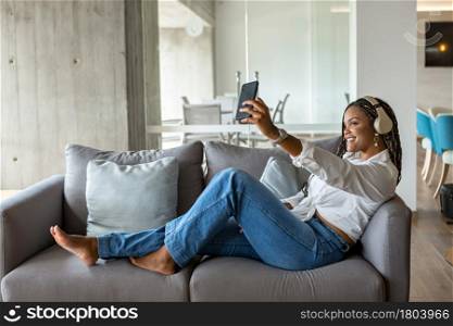 Portrait of a happy young woman listening to music with headphones and taking a selfie while leaning on a sofa at home. Concept of people in home.. Happy young woman listening to music with headphones and using mobile phone to take a selfie while leaning on a sofa at home