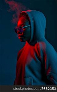 Portrait of a hipster woman in sunglasses and sweater hood over dark studio background in blue neon light. Casual hipster woman in sunglasses over dark studio wall