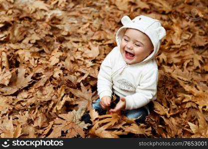 Portrait of a little baby boy laughing, sitting on the ground covered with dry tree leaves in the forest, enjoying autumn nature