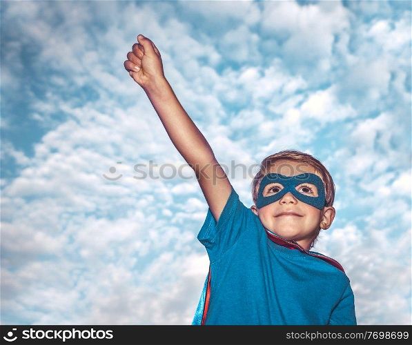 Portrait of a little boy wearing costume of superhero with  raised up one hand over sky background, is preparing to save the world, kid plays superman