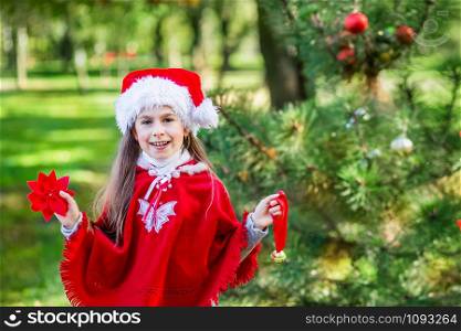 Portrait of a little girl in a Santa Claus hat near a Christmas tree in the forest. Girl decorates a Christmas tree in the forest with Christmas balls. Merry Christmas and happy holidays.. Portrait of a little girl in a Santa Claus hat near a Christmas tree in the forest.