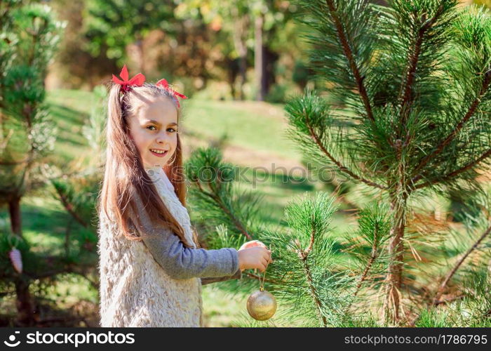 Portrait of a little girl near the Christmas tree. Girl decorates a Christmas tree in the forest with Christmas balls. Winter holidays and people concept. Merry Christmas and happy holidays.. Portrait of a little girl near the Christmas tree. Girl decorates a Christmas tree in the forest with Christmas balls.