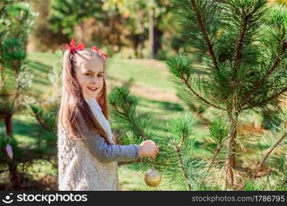 Portrait of a little girl near the Christmas tree. Girl decorates a Christmas tree in the forest with Christmas balls. Winter holidays and people concept. Merry Christmas and happy holidays.. Portrait of a little girl near the Christmas tree. Girl decorates a Christmas tree in the forest with Christmas balls.