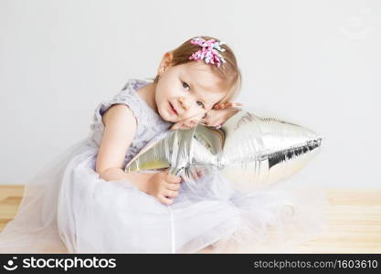 Portrait of a lovely little girl in elegant gray dress in front of a white background. Little princess. Little baby girl playing with silver star-shaped balloon.