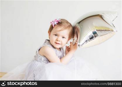 Portrait of a lovely little girl wearing elegant gray dress in front of a white background. Little princess. Little baby girl holding silver star-shaped balloon.
