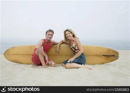 Portrait of a mature couple sitting on the beach with a surfboard