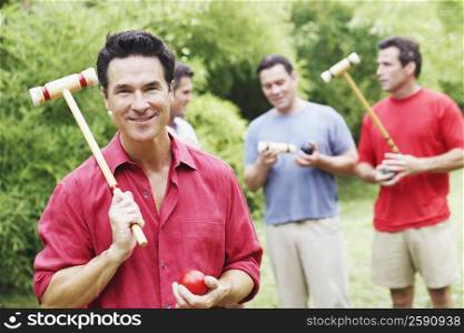 Portrait of a mature man holding a croquet mallet and a ball with his friends in the background