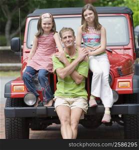 Portrait of a mature man holding hands of his two daughters and sitting on the bonnet of a jeep