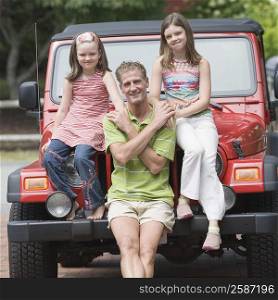 Portrait of a mature man holding hands of his two daughters and sitting on the bonnet of a jeep