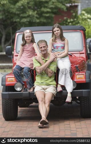 Portrait of a mature man holding hands of his two daughters sitting on the bonnet of a jeep