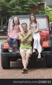Portrait of a mature man holding hands of his two daughters sitting on the bonnet of a jeep
